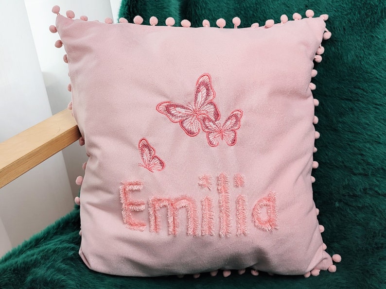 K&ouml;nnte beinhalten: Rosafarbenes Kissen mit Pompon-Besatz und gestickten Schmetterlingen sowie dem Namen "Emilia".