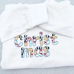 Peut inclure: Sweat-shirt blanc avec le mot "Christmas" brodé en lettres patchwork colorées. Les lettres présentent des motifs comprenant des zigzags, des arbres et des nœuds. Le sweat-shirt est plié, avec la capuche visible.