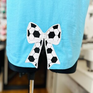 May include: A light blue t-shirt with a black and white soccer ball patterned bow.