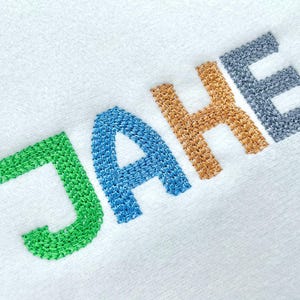 May include: White fabric with the embroidered name "JAKE" in green, blue, gold, and gray thread. The letters are in a sans-serif font and are slightly angled across the fabric.
