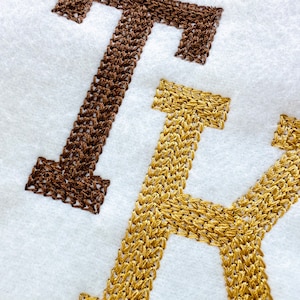 Può includere: Due lettere ricamate, "T" e "K", cucite con filo marrone e dorato su uno sfondo di tessuto bianco.