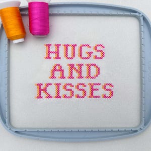 May include: A cross-stitch design in a blue embroidery hoop, with the words "HUGS AND KISSES" stitched in pink and orange thread. Two thread spools, one orange and one pink, are positioned above the design.