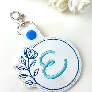 May include: White leather keychain with a blue button and a blue and white embroidered floral design with the letter E in the center.