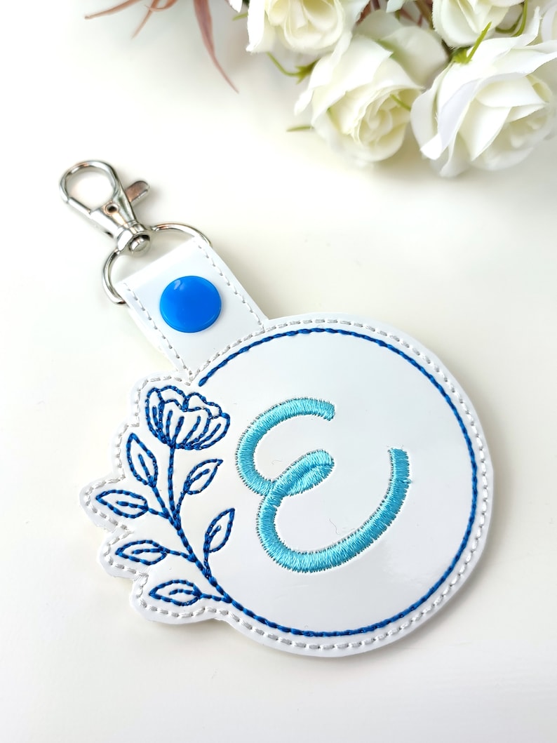May include: White leather keychain with a blue button and a blue and white embroidered floral design with the letter E in the center.