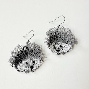 Puede incluir: Un par de pendientes de erizo hechos a mano. Cada pendiente presenta un diseño de erizo bordado detallado con una combinación de colores gris y blanco. Los pendientes tienen alambres para las orejas de gancho de pez de color plateado.