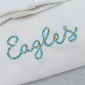 Puede incluir: Tela blanca con la palabra "Eagles" bordada en una fuente cursiva. El bordado est&aacute; delineado en azul claro con un relleno dorado. En la esquina superior izquierda se ve una tela blanca doblada con un borde acanalado.