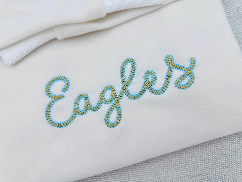 Puede incluir: Tela blanca con la palabra "Eagles" bordada en una fuente cursiva. El bordado est&aacute; delineado en azul claro con un relleno dorado. En la esquina superior izquierda se ve una tela blanca doblada con un borde acanalado.