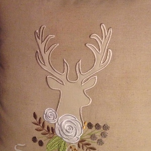 May include: A beige pillow with an embroidered white deer head and floral design. The floral design includes white roses, green leaves, and brown berries. The text "Seaux Cajunique" is embroidered at the bottom of the pillow.