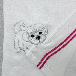 May include: White kitchen towel featuring an embroidered fluffy white dog. The towel has a red stripe detail on the edge. The dog embroidery has black eyes and nose.