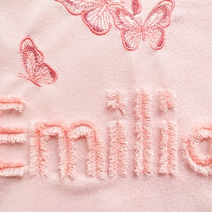 K&ouml;nnte beinhalten: Rosafarbener Stoff mit dem Namen "Emilia", der in einem flauschigen rosa Garn gestickt ist. Der Name ist von rosa gestickten Schmetterlingen umgeben.