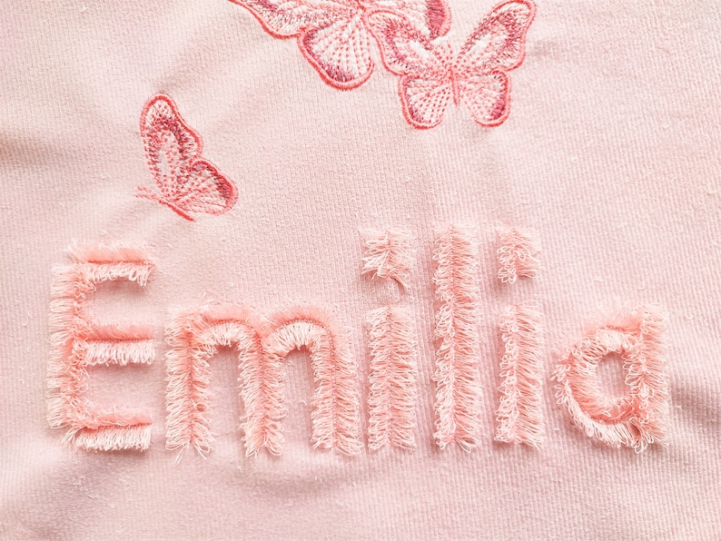 K&ouml;nnte beinhalten: Rosafarbener Stoff mit dem Namen "Emilia", der in einem flauschigen rosa Garn gestickt ist. Der Name ist von rosa gestickten Schmetterlingen umgeben.