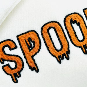 May include: Close-up of a white fabric with the embroidered word "SPOOKY" in orange with black outlines. The letters have a dripping effect, creating a spooky Halloween theme.