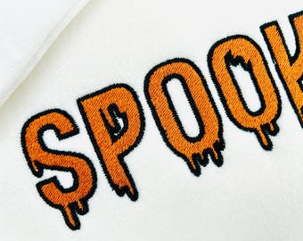 May include: Close-up of a white fabric with the embroidered word "SPOOKY" in orange with black outlines. The letters have a dripping effect, creating a spooky Halloween theme.