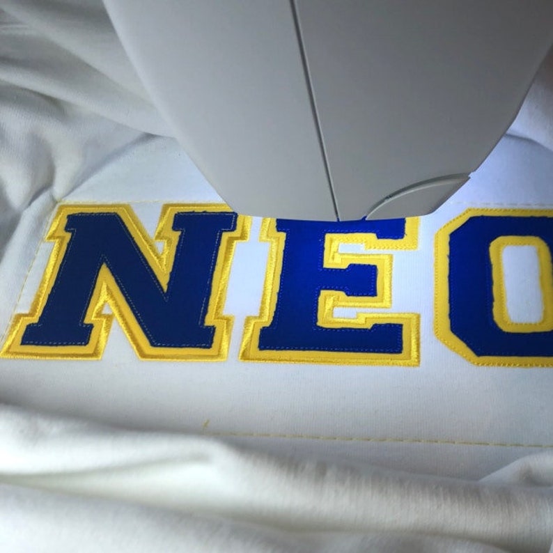 May include: A close-up of a white fabric with the word "NEO" embroidered in blue and yellow thread.