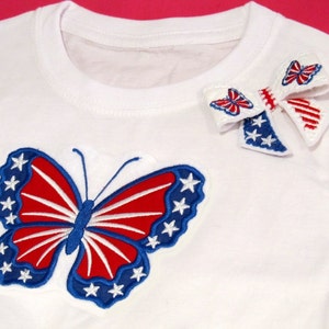 May include: White t-shirt with a red, white, and blue butterfly applique and a red, white, and blue bow applique.