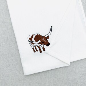 May include: White folded kitchen towel with an embroidered longhorn steer design. The steer is brown and white with long curved horns and black details. The towel is made of a soft, white fabric.