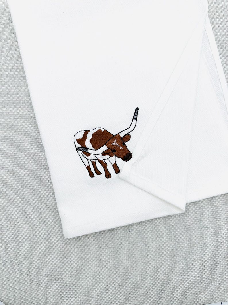 May include: White folded kitchen towel with an embroidered longhorn steer design. The steer is brown and white with long curved horns and black details. The towel is made of a soft, white fabric.