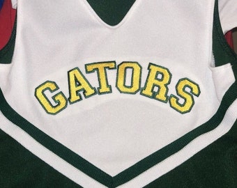 May include: White cheerleading uniform with green and white stripes and the word "GATORS" embroidered in yellow on the front.