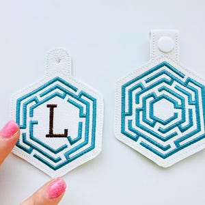 Puede incluir: Dos llaveros de polipiel blanca con un diseño de laberinto turquesa. Un llavero tiene la letra "L" bordada en marrón. Los llaveros son hexagonales y tienen un pequeño lazo con cierre de botón a presión.