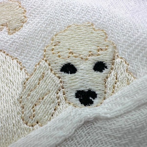 May include: Close-up of a white textile featuring an embroidered poodle design. The poodle is cream-coloured with black eyes and nose, and the embroidery is outlined in a light tan thread. The background is a textured white fabric.