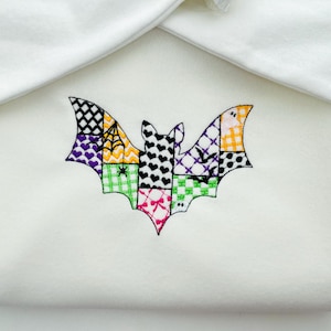 May include: A white baby blanket with a colorful embroidered bat design. The bat is segmented into various patterns, including hearts, spiderwebs, and checkered squares, in shades of purple, orange, green, and pink. The blanket is folded, with a white trim.