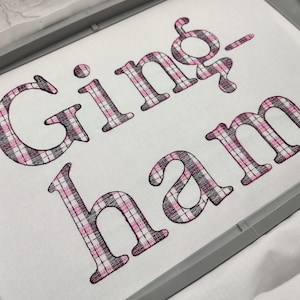 Pode incluir: Tecido vichy com o texto "Gingham" bordado em fio preto e rosa.