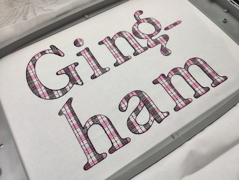 Pode incluir: Tecido vichy com o texto "Gingham" bordado em fio preto e rosa.