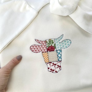 May include: White hooded sweatshirt with a patchwork cow design embroidered on the front. The cow is made of different coloured fabric squares, including red, pink, blue, green, yellow, and white.