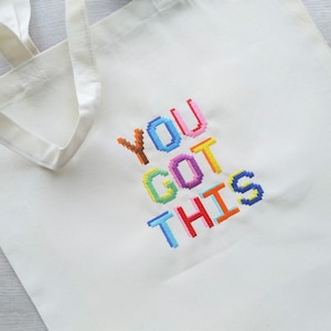 May include: White canvas tote bag with the phrase "YOU GOT THIS" embroidered in colorful pixelated letters.