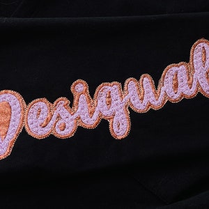 Op de afbeelding: Een zwart kledingstuk met het woord "Desigual" geborduurd in roze en bruine letters.