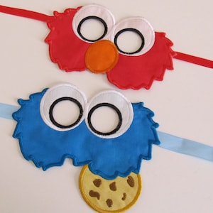 Może przedstawiać: Dwie filcowe maski postaci z przymocowanymi wstążkami. Czerwona maska ma puszystą fakturę i posiada duży pomarańczowy nos i białe oczy. Niebieska maska ma puszystą fakturę i posiada ciasteczko z kawałkami czekolady.