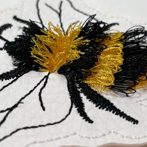Op de afbeelding: Een witte vilt appliqué van een hommel met zwarte en gouden borduurwerk. De bij is omlijnd met zwarte draad en heeft een pluche textuur.