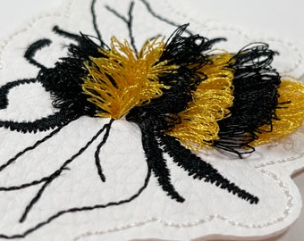 Op de afbeelding: Een witte vilt appliqué van een hommel met zwarte en gouden borduurwerk. De bij is omlijnd met zwarte draad en heeft een pluche textuur.