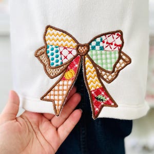 May include: Embroidered bow applique with a patchwork design in various colors, including brown, yellow, red, green, and blue. The bow is sewn onto a white fabric, creating a decorative element.