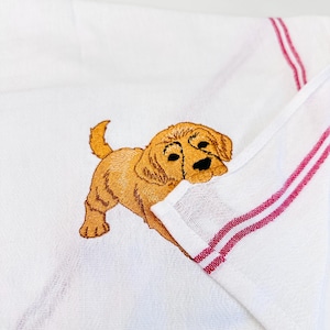 May include: White kitchen towel featuring an embroidered golden retriever puppy. The towel has two burgundy stripes along the edge. The puppy is light brown with black eyes and nose.