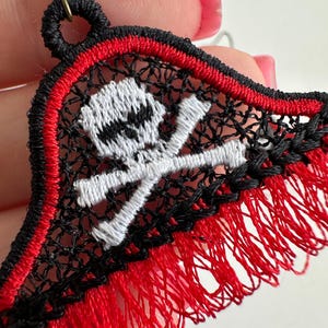 May include: Close-up of a pirate-themed earring. The earring features a black, red, and white embroidered design of a skull and crossbones. The edges are trimmed with red fringe, and the earring has a bronze-colored hook.