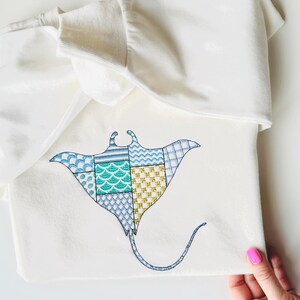 Könnte beinhalten: Cremefarbenes T-Shirt mit einem gestickten Mantarochen-Design. Der Mantarochen besteht aus verschiedenen gemusterten Patches in Blau-, Türkis- und Goldtönen. Das T-Shirt ist gefaltet, das Design ist auf der Vorderseite sichtbar.