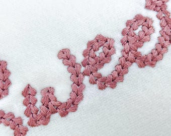 May include: Close-up of white fabric with the embroidered word "love" in a dusty rose color. The embroidery has a textured, woven appearance, with each letter carefully stitched. The background is a plain white, providing a clean contrast.