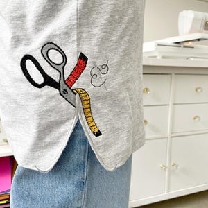 May include: A light gray t-shirt featuring an embroidered design of black scissors with a red and yellow measuring tape wrapped around the blades, accompanied by a small black thread and needle icon near the side hem of the garment.