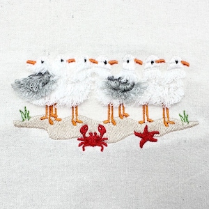 Puede incluir: Diseño bordado con seis gaviotas blancas con picos y patas naranjas, de pie en una orilla arenosa. También se borda un cangrejo y una estrella de mar rojos. El diseño está sobre un fondo de tela blanca.