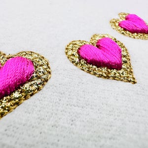 Könnte beinhalten: Nahaufnahme von drei gestickten Herzen auf weißem Stoff. Die Herzen haben eine leuchtend pinke Mitte und sind mit goldenem Glitzer umrandet. Die Herzen sind unterschiedlich groß und diagonal angeordnet.
