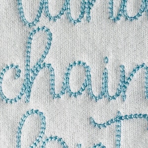 May include: Close-up of a white fabric with the words "chain" and "stitch" embroidered in light blue thread.