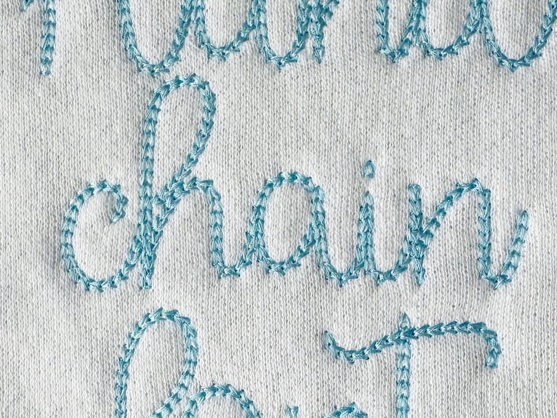 May include: Close-up of a white fabric with the words "chain" and "stitch" embroidered in light blue thread.
