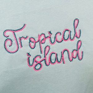 May include: Embroidered design on a white fabric background. The design says "Tropical Island" in a cursive font. The letters are outlined in teal and filled in with pink.
