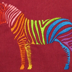 Op de afbeelding: Een regenboogzebra geborduurd op een maronkleurige stof. De zebra is omlijnd met roze, oranje, geel, groen, blauw en paars.