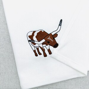May include: White cloth with an embroidered image of a brown and white longhorn steer. The steer is depicted in a detailed, cartoon-like style. The cloth has a subtle herringbone pattern.