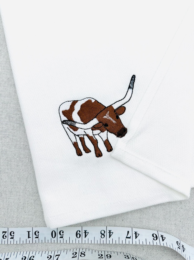May include: White cloth with an embroidered image of a brown and white longhorn steer. The steer is depicted in a detailed, cartoon-like style. The cloth has a subtle herringbone pattern.