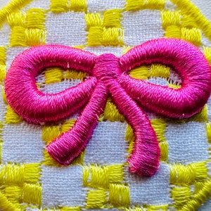 Może przedstawiać: Różowa kokardka z tkaniny na żółto-białym tle w kratkę. Kokardka wykonana jest z miękkiej, fakturowanej tkaniny i ma prosty, elegancki design.