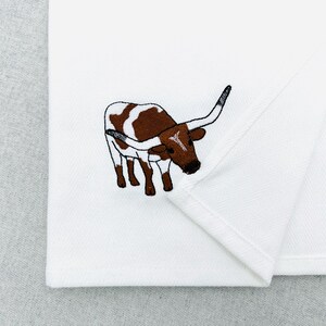 May include: White folded fabric with an embroidered image of a brown and white longhorn steer. The steer has long horns and is facing left. The embroidery is detailed and the fabric is clean.