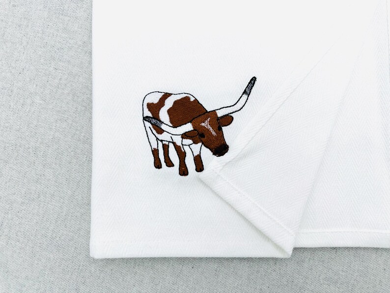 May include: White folded fabric with an embroidered image of a brown and white longhorn steer. The steer has long horns and is facing left. The embroidery is detailed and the fabric is clean.
